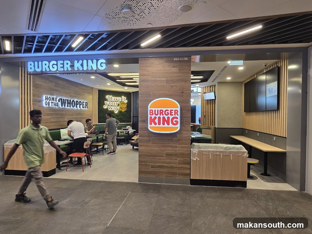 Photo of Burger King storefront with visible signage including 'BURGER KING' neon sign above entrance, 'HOME OF THE WHOPPER', and 'Have it your way'. Interior shows seating area with green sofas, tables, and staff members. A person walks past the entrance.