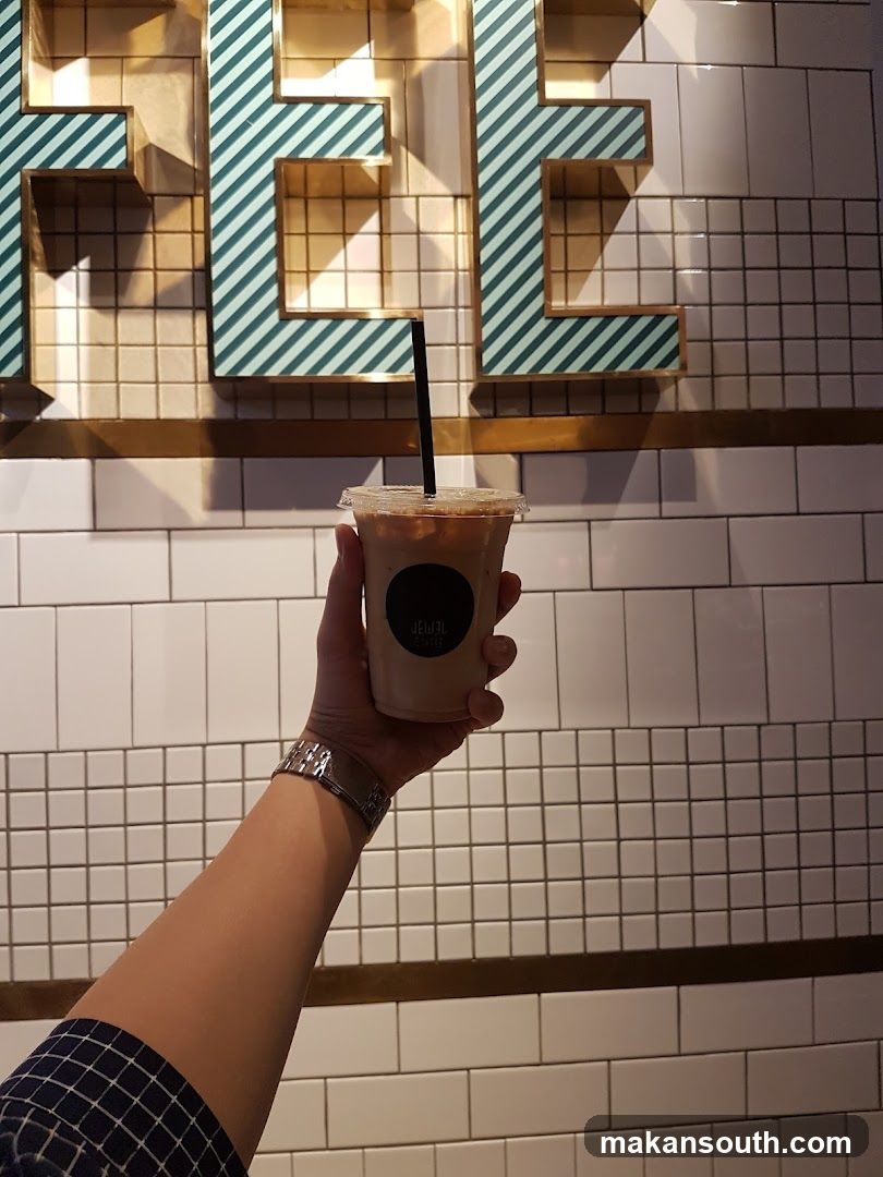 Hand holding iced coffee in front of tiled wall with large letter signage