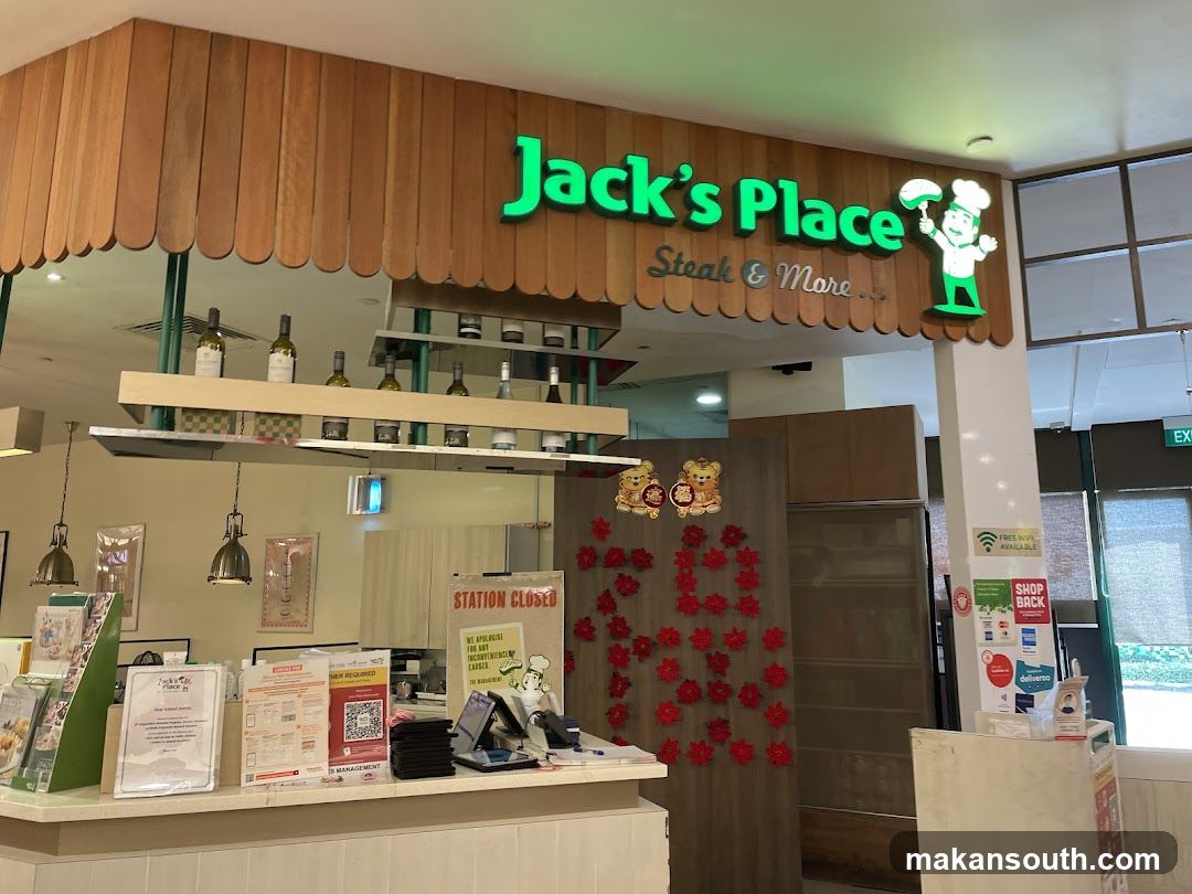 The exterior of a restaurant called Jack's Place, which specializes in steak and more. The sign is illuminated green and features a cartoon chef. Wooden paneling is visible above the entrance. Inside, bottles of wine are displayed on shelves. A counter area is visible with various signs and promotional materials.