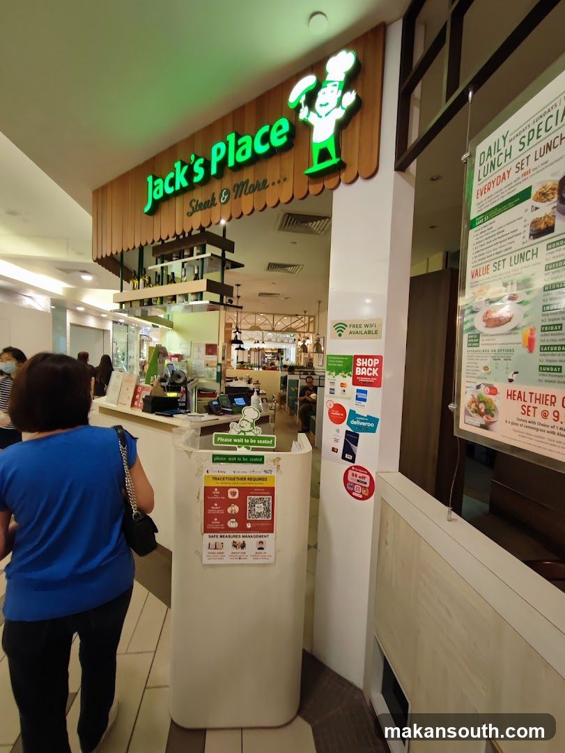 The exterior of a Jack's Place restaurant. A sign above the entrance reads "Jack's Place Steak & More...". To the right, a menu board displays daily lunch specials and set lunches. A sign indicates "FREE WIFI AVAILABLE". There are also signs for delivery services and a "$5 off" promotion. A notice about "TRACETOGETHER REQUIRED" and "SAFE MEASURES MANAGEMENT" is posted on a white stand.
