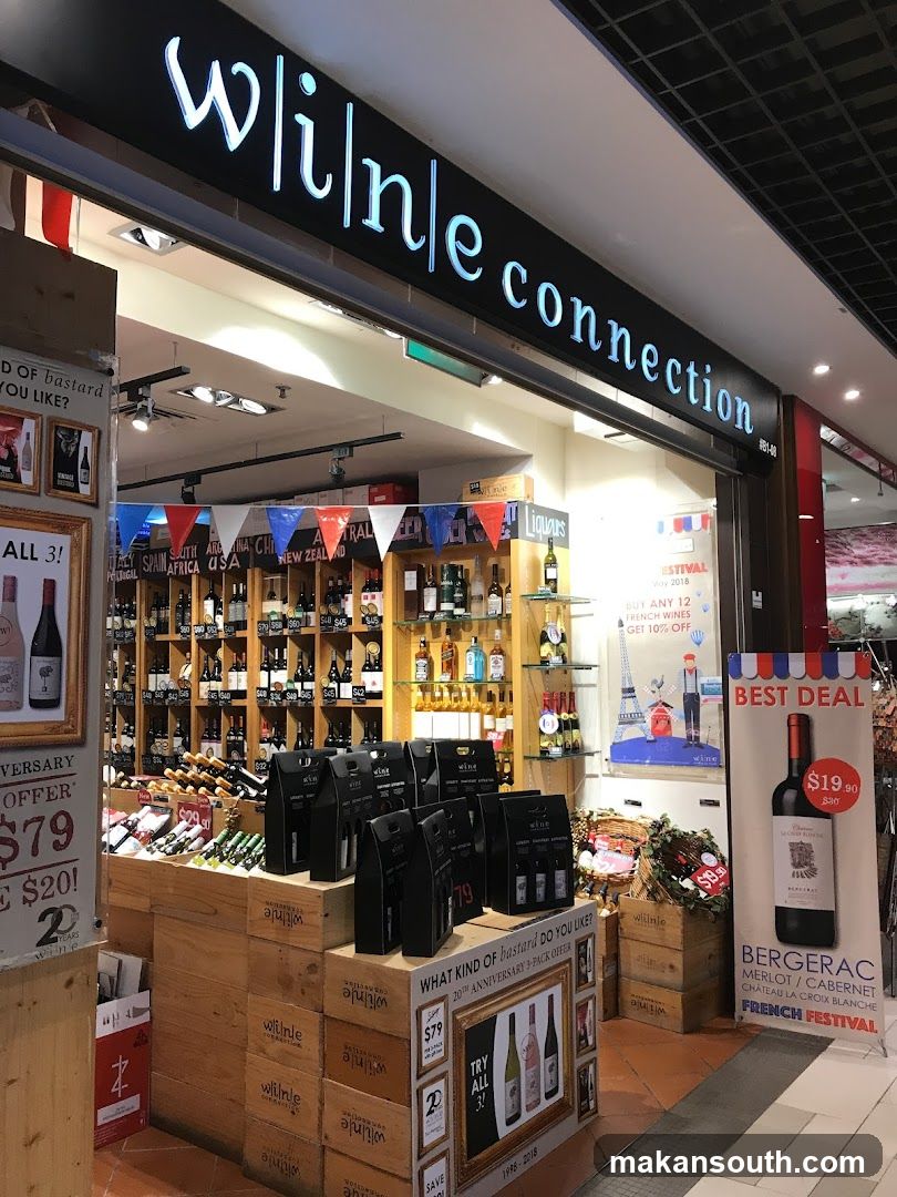The exterior of a wine shop called "Wine Connection". The storefront features a large illuminated sign with the store's name. Inside, shelves are stocked with numerous bottles of wine and spirits. Displays showcase special offers and gift packs, including a "20th Anniversary 3-Pack Offer" and a "French Festival" promotion featuring Bergerac wine.