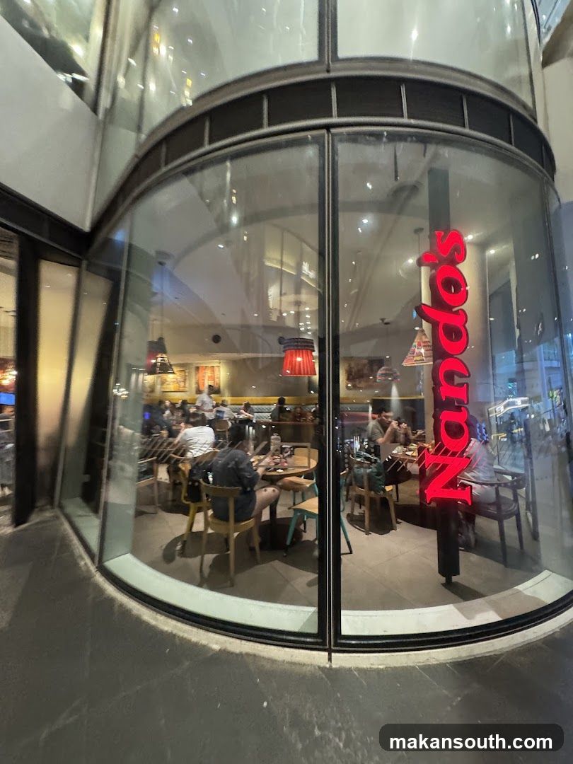 A view of the interior of a Nando's restaurant through a large, curved glass window. The red Nando's logo is visible vertically on a pillar inside. Several patrons are seated at tables, some eating and others conversing. The restaurant appears to be well-lit with pendant lights hanging from the ceiling.