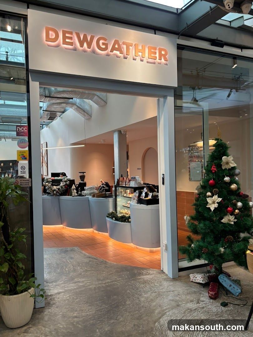 The entrance to a shop with the name "DEWGATHER" illuminated above the doorway. Inside, a counter area is visible with display cases and what appears to be a coffee machine. A Christmas tree decorated with ornaments stands to the right of the entrance, with wrapped gifts underneath.