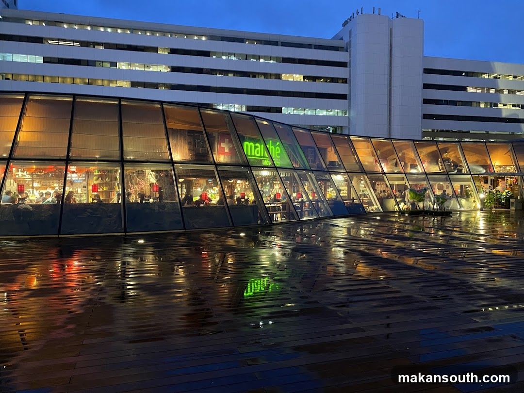 A view of the exterior of a Marche restaurant at dusk. The building has a curved glass facade, revealing a warmly lit interior with diners. The Marche logo is visible on the glass. The wet wooden deck in front of the restaurant reflects the lights from the building and the sky.