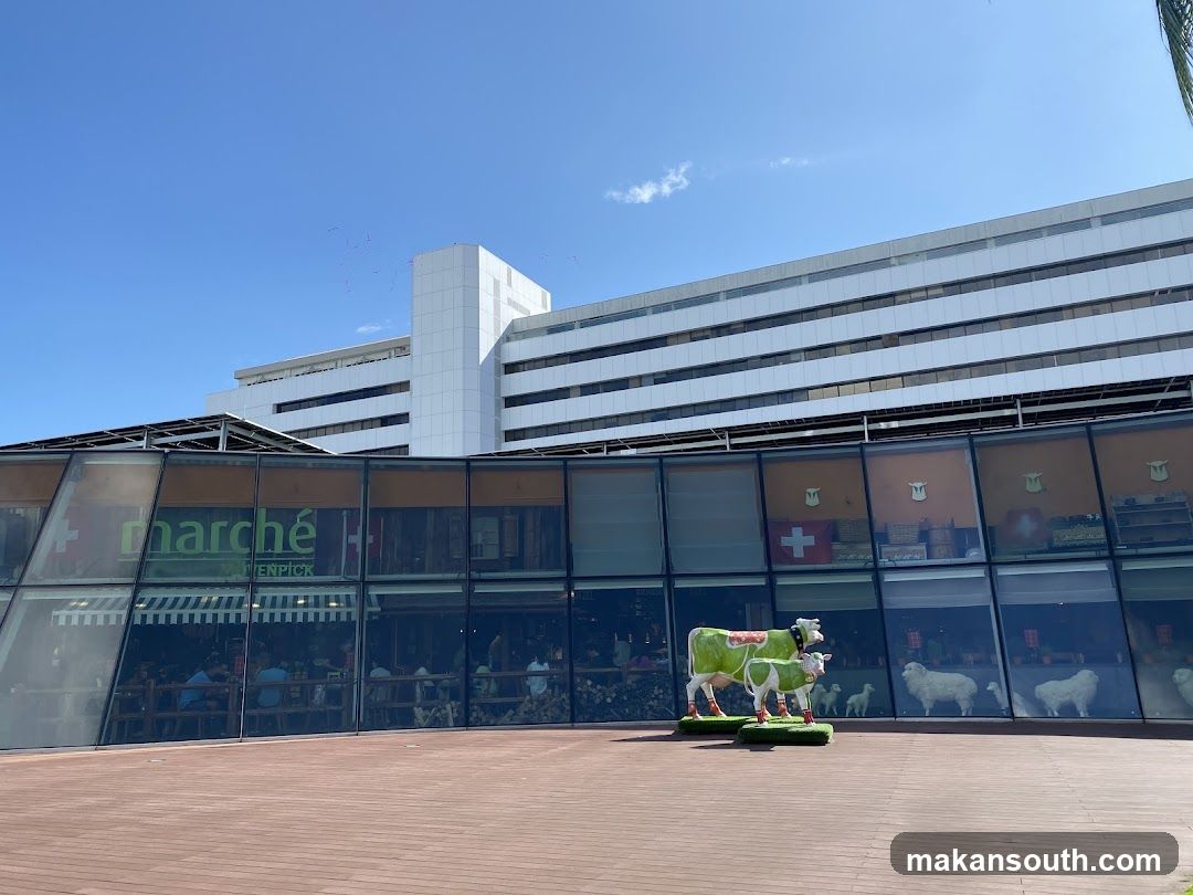 A modern building with a glass facade. The signage for "marché Mövenpick" is visible on the glass. Inside, people are seated at tables. Outside, there are decorative sculptures of cows and sheep.