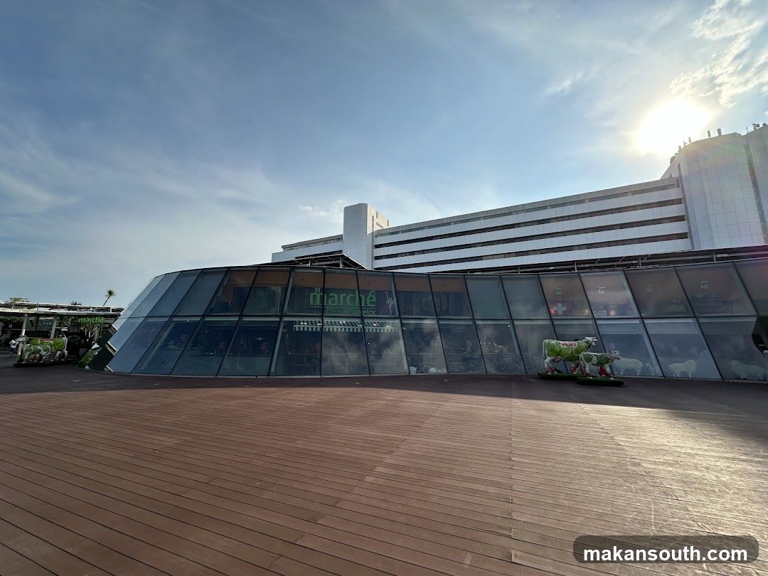 A modern, curved glass building with the "marche" logo visible. The building is set against a bright sky with the sun shining. Decorative cow statues are placed outside the building.