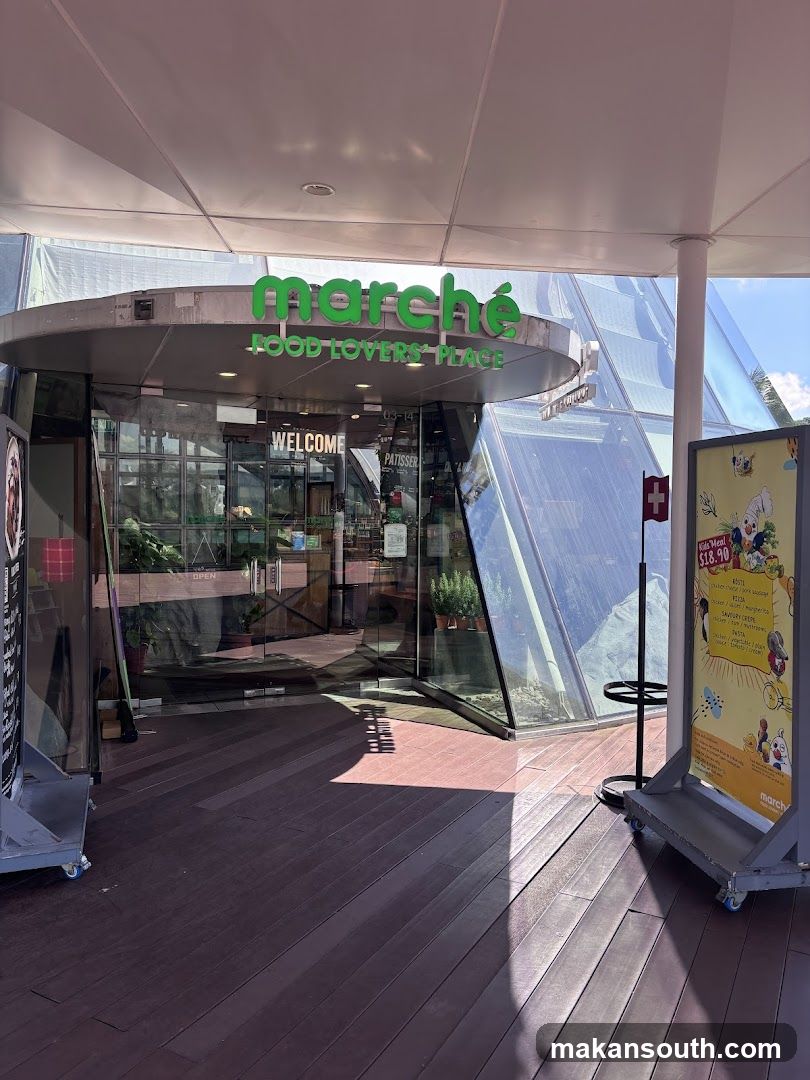 The exterior of a Marché Food Lovers Place entrance with a large glass facade and a prominent green sign above. The entrance is marked with a "WELCOME" sign. To the right, a promotional stand displays a menu with cartoon illustrations.
