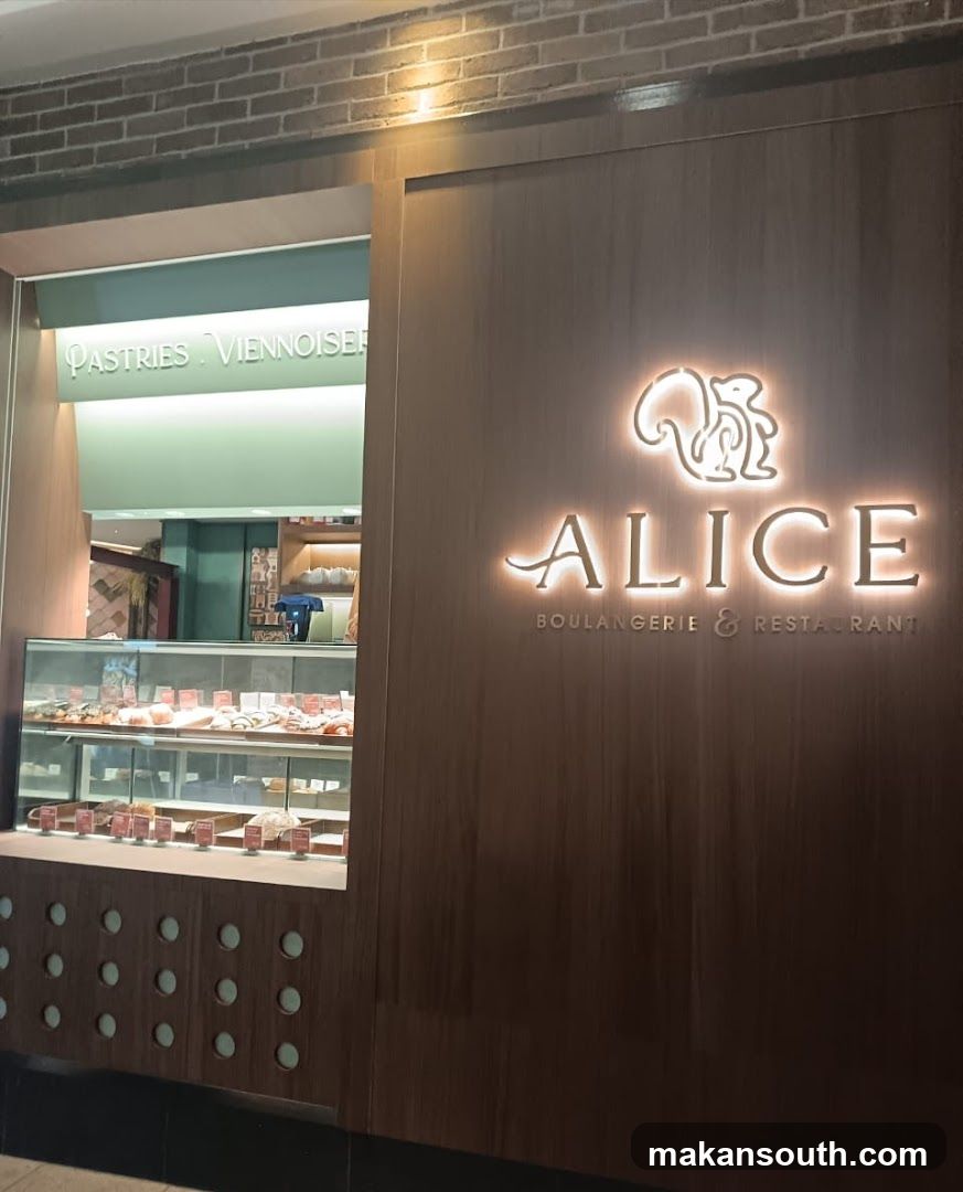 The exterior of a bakery and restaurant with a wooden facade. The illuminated logo features a squirrel and the name "ALICE" with "BOULANGERIE & RESTAURANT" below it. To the left, a display case filled with pastries is visible, and a sign above reads "PASTRIES VIENNOISERIE".