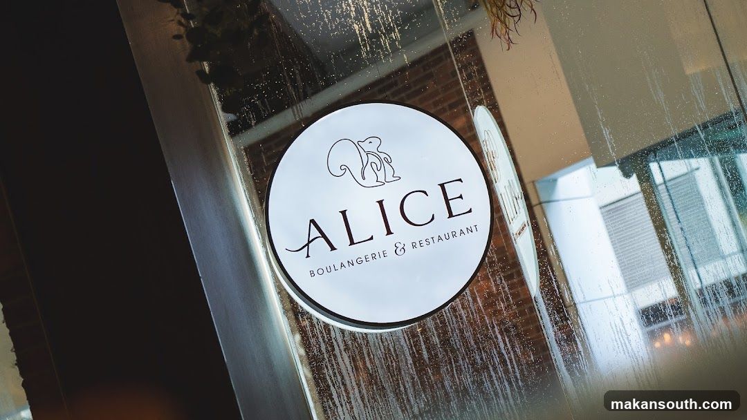 A circular sign with a stylized squirrel illustration above the word "ALICE" and "BOULANGERIE & RESTAURANT" below it. The sign is affixed to a glass surface, possibly a storefront window or door. The glass has water droplets or condensation on it, and reflections of the interior and exterior are visible.
