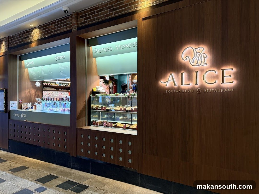 The exterior of a bakery and restaurant with a prominent illuminated sign that reads "ALICE BOULANGERIE & RESTAURANT" with a squirrel logo above. To the left, there are display windows showcasing pastries and drinks. One window has a menu board for coffee and drinks, and another displays pastries in a refrigerated case.