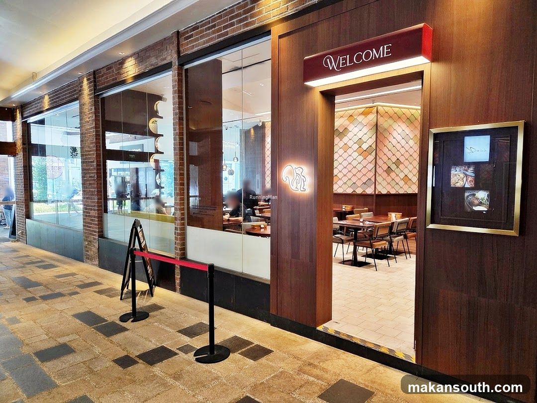 The exterior of a restaurant with a wooden facade and a glass entrance. A red velvet rope barrier is set up outside the entrance. Above the doorway, a lit sign reads "WELCOME". To the left of the entrance, a neon squirrel logo is visible. Inside, tables and chairs are set up, and the walls are decorated with a patterned mosaic. A framed picture hangs on the right side of the entrance, displaying what appears to be food items.
