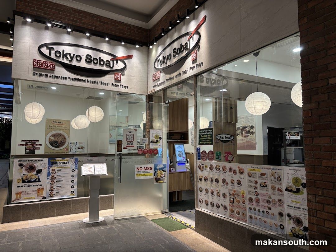 The exterior of a Tokyo Soba restaurant. The storefront features large glass windows displaying menus and the restaurant's name and logo prominently displayed above the entrance. White paper lanterns hang inside the restaurant.