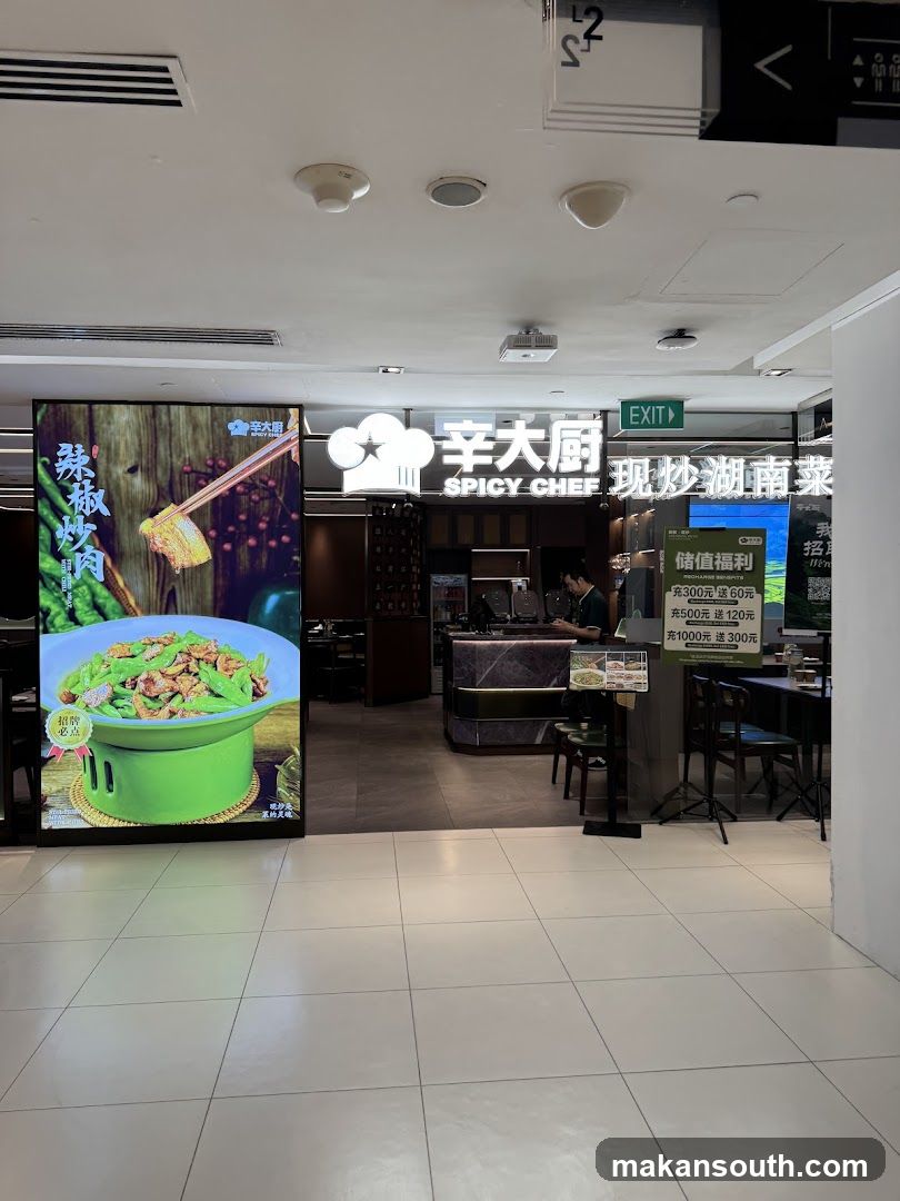 The entrance to a restaurant called Spicy Chef, which specializes in Hunan cuisine. A large digital display on the left shows a dish of stir-fried meat and green beans. The restaurant's name and logo are visible above the entrance. An exit sign is also present. A promotional poster with discount offers is displayed near the entrance.