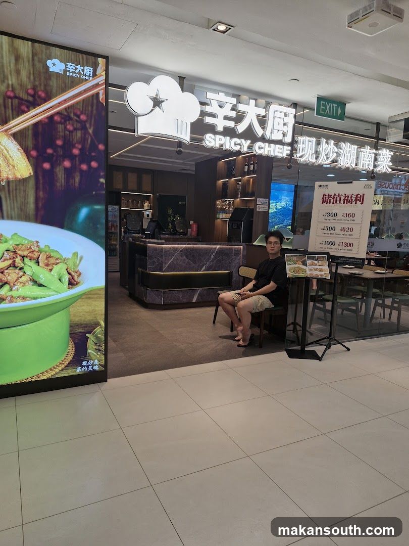 The exterior of a restaurant called Spicy Chef. A person is sitting on a chair outside the restaurant. There is a large digital display on the left showing food. A menu board is visible on the right.