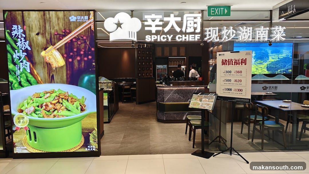 The exterior of a restaurant called Spicy Chef. A large digital display on the left shows a dish of stir-fried meat with chili and green beans. The restaurant's name and logo are visible above the entrance. An "EXIT" sign is also present. Inside, staff can be seen behind a counter. A promotional sign detailing recharge benefits is displayed near the entrance.