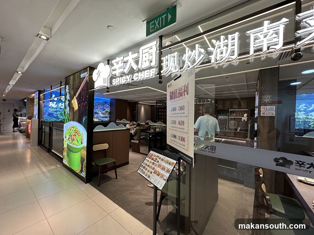The exterior of a restaurant called Spicy Chef. The restaurant has a large illuminated sign with the name in Chinese and English. There are large windows showing the interior dining area. A menu board with promotional benefits is displayed near the entrance.