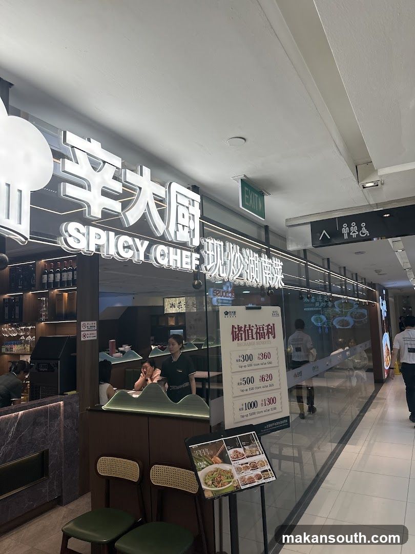 The exterior of a restaurant called Spicy Chef. The signage is illuminated and prominent. Inside, staff members are visible, and there are tables and chairs. A promotional poster detailing recharge benefits is displayed outside the entrance.
