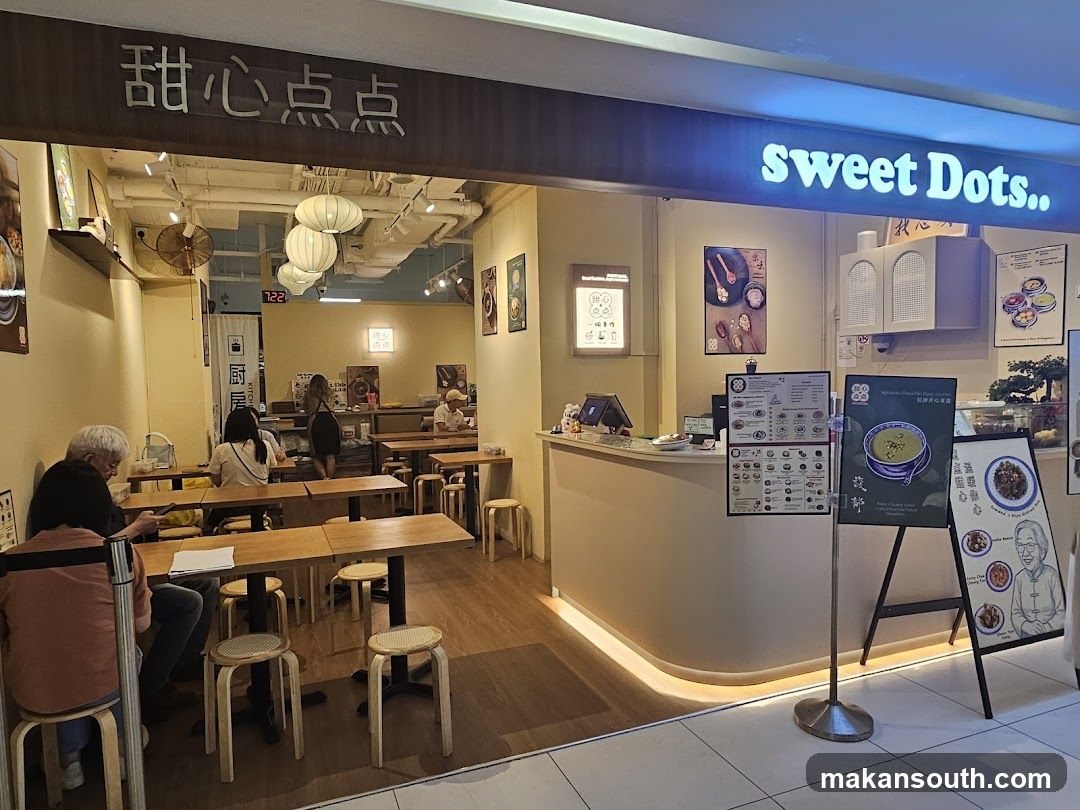 The exterior of a dessert shop with a prominent "sweet Dots.." sign. The shop has a clean, modern interior with wooden tables and stools. Several customers are seated at tables, and staff can be seen behind the counter. There are also menu boards and promotional posters displayed.