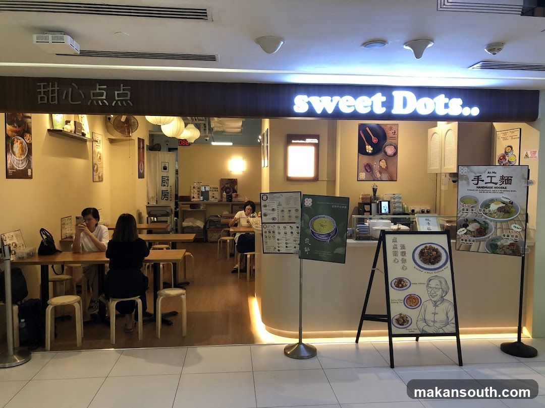 The exterior of a dessert shop called Sweet Dots. The shop has a clean and modern interior with wooden tables and chairs. There are several posters and menus displayed outside the shop, showcasing various food items.