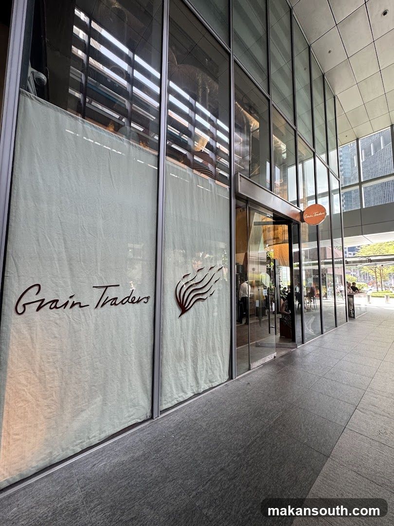 A modern building facade with large glass windows and doors. The name "Grain Traders" is visible in dark lettering on a light-colored curtain covering a large portion of the window. A stylized logo resembling flames or waves is next to the name. A circular orange sign with the same name is above the entrance.