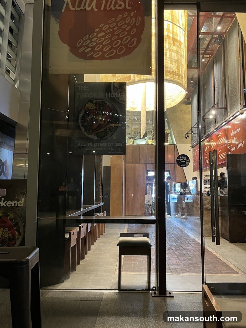 View through a glass storefront into a restaurant. A long counter with stools lines the interior. A sign advertises "The Trader Hour" with "All Plates at $15". The interior features warm lighting and brick flooring.