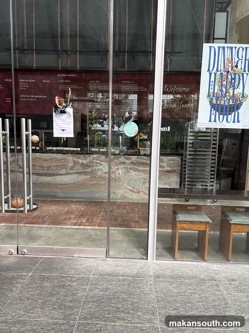 The storefront of Grain Traders features large glass windows and doors. Inside, a menu board is visible with various food items and prices. A poster advertising "Dinner Happy Hour" is displayed on the right side of the entrance. Two wooden stools are placed near a counter inside.