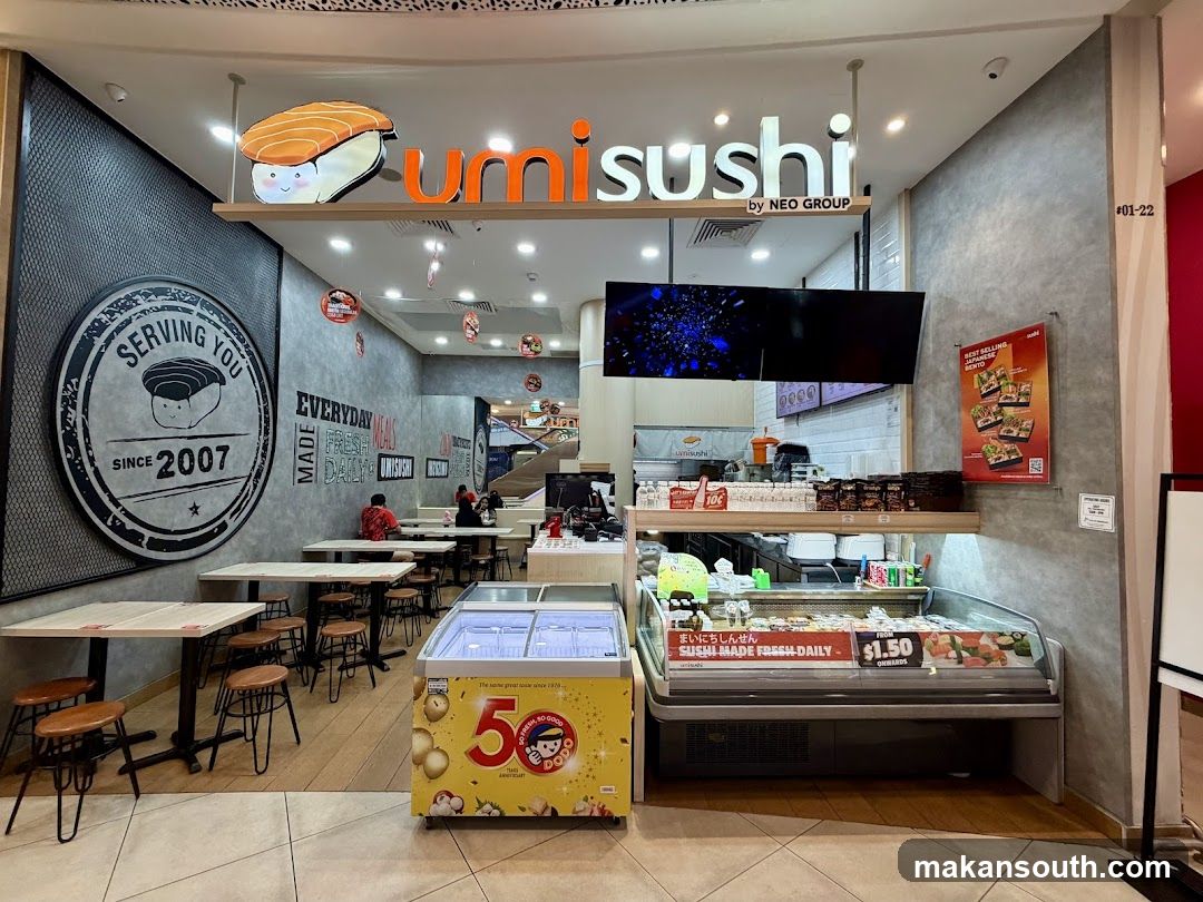 The exterior of a Umi Sushi restaurant with its prominent signage and a display of fresh sushi. The restaurant has a modern industrial design with concrete walls and metal accents. Seating areas with tables and stools are visible, as well as a refrigerated display case filled with various sushi options. A large circular sign on the wall indicates "Serving You Since 2007".