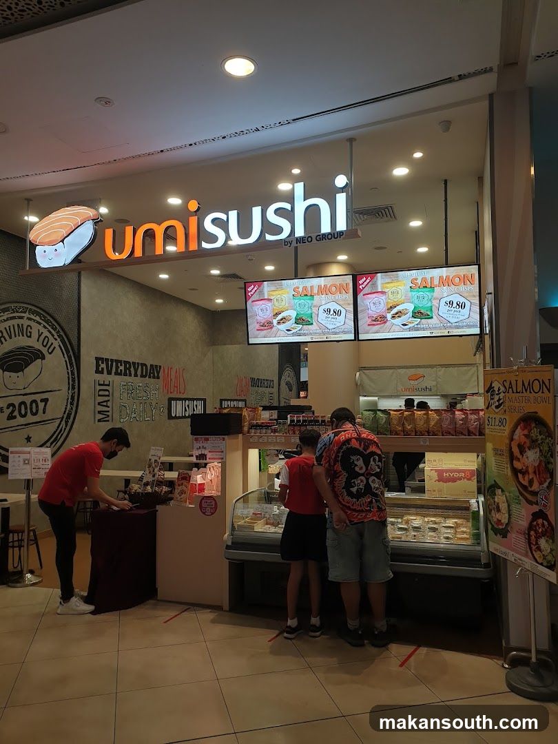 The exterior of a Umi Sushi restaurant with its prominent signage. The sign features the Umi Sushi logo and "by NEO GROUP". Below the sign, digital screens display promotions for salmon products. The storefront includes a display case with food items and a counter where staff interact with customers. A promotional poster for a salmon master bowl series is visible on the right.