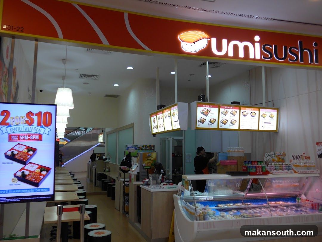 The storefront of a sushi restaurant called Umi Sushi. The restaurant has a bright orange sign with the restaurant's name and logo. There are menu boards hanging above the counter, displaying various bento boxes and sushi dishes. A digital display on the left advertises a "2 FOR $10 Bento Twin Deal" available daily from 5 PM to 8 PM. The interior shows a counter with food displays and seating areas.