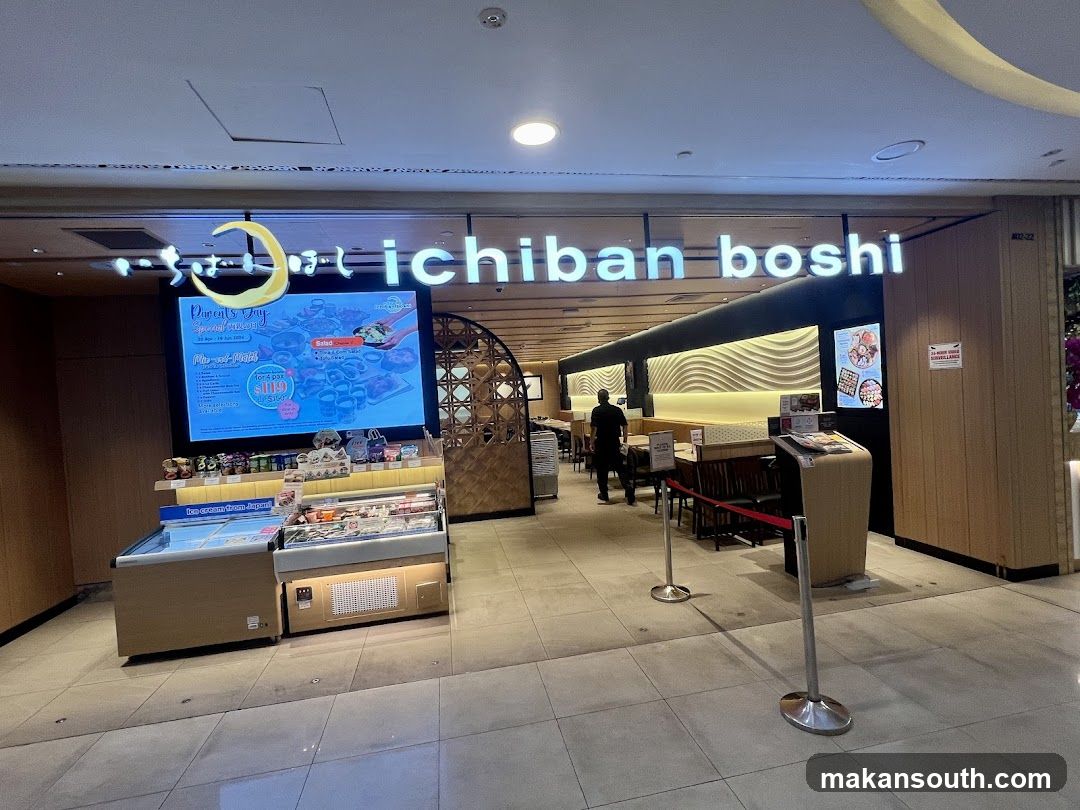 The exterior of a Japanese restaurant with the name "Ichiban Boshi" displayed prominently on a lit sign above the entrance. To the left, a large digital screen advertises a "Parents' Day Special" with various food items and a price of $119. Below the screen is a display case with refrigerated items, labeled "Ice cream from Japan." The entrance features a curved wooden archway, and inside, tables and seating are visible. A staff member is seen walking inside. Red velvet ropes guide customers towards the entrance.