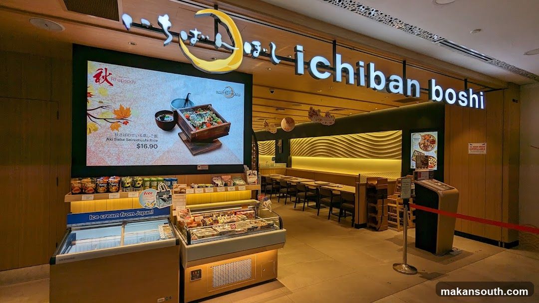 The exterior of a Japanese restaurant with a large digital display showing a seasonal dish and the restaurant's name, "ichiban boshi," prominently displayed above the entrance. In front of the display, there are refrigerated units selling ice cream and shelves stocked with various food items. The interior of the restaurant, with tables and chairs, is visible through the entrance.