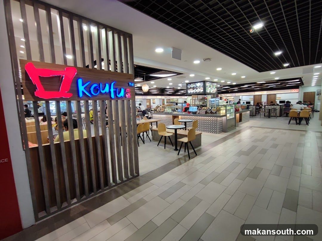 A view of the Koufu food court entrance with its prominent signage. The food court appears to be busy with customers seated at tables and ordering food. The interior features a modern design with a black grid ceiling and tiled flooring.