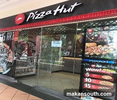 The exterior of a Pizza Hut restaurant with its prominent signage. The entrance is visible, leading into a dining area with tables and chairs. A display stand with promotional material is placed near the entrance.
