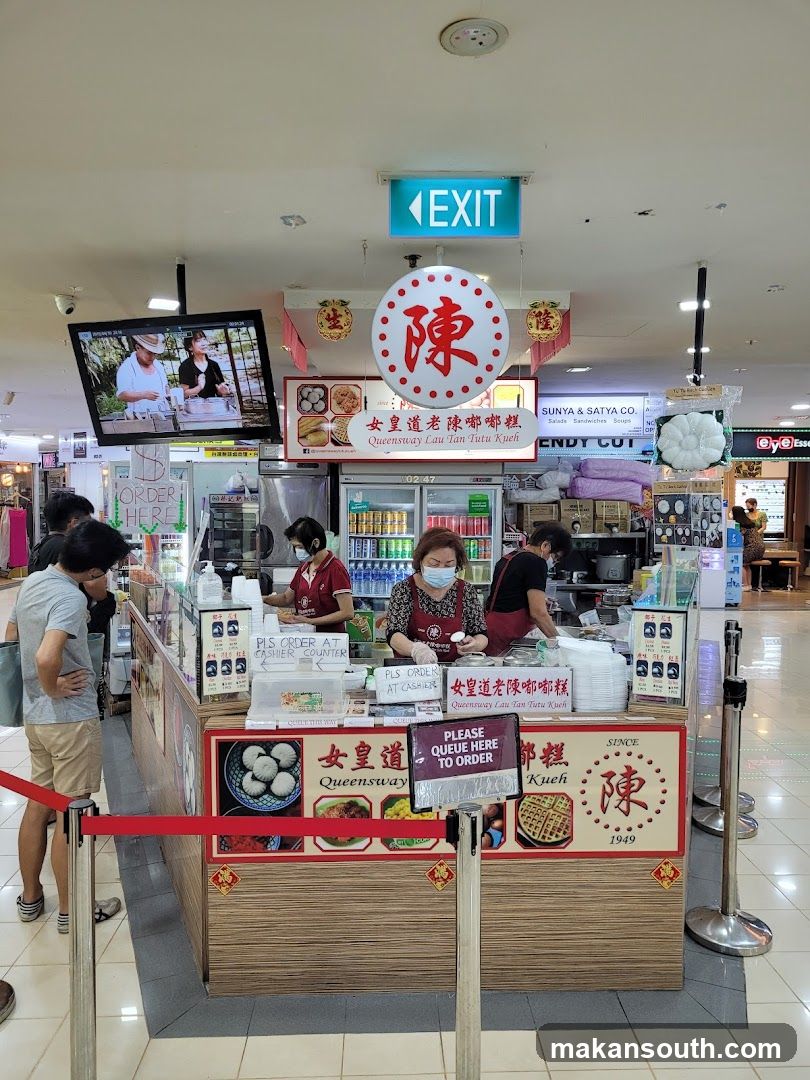 A food stall in a mall selling Tutu Kueh. There are staff members serving customers. A television screen shows a cooking demonstration. Red velvet ropes guide the queue.