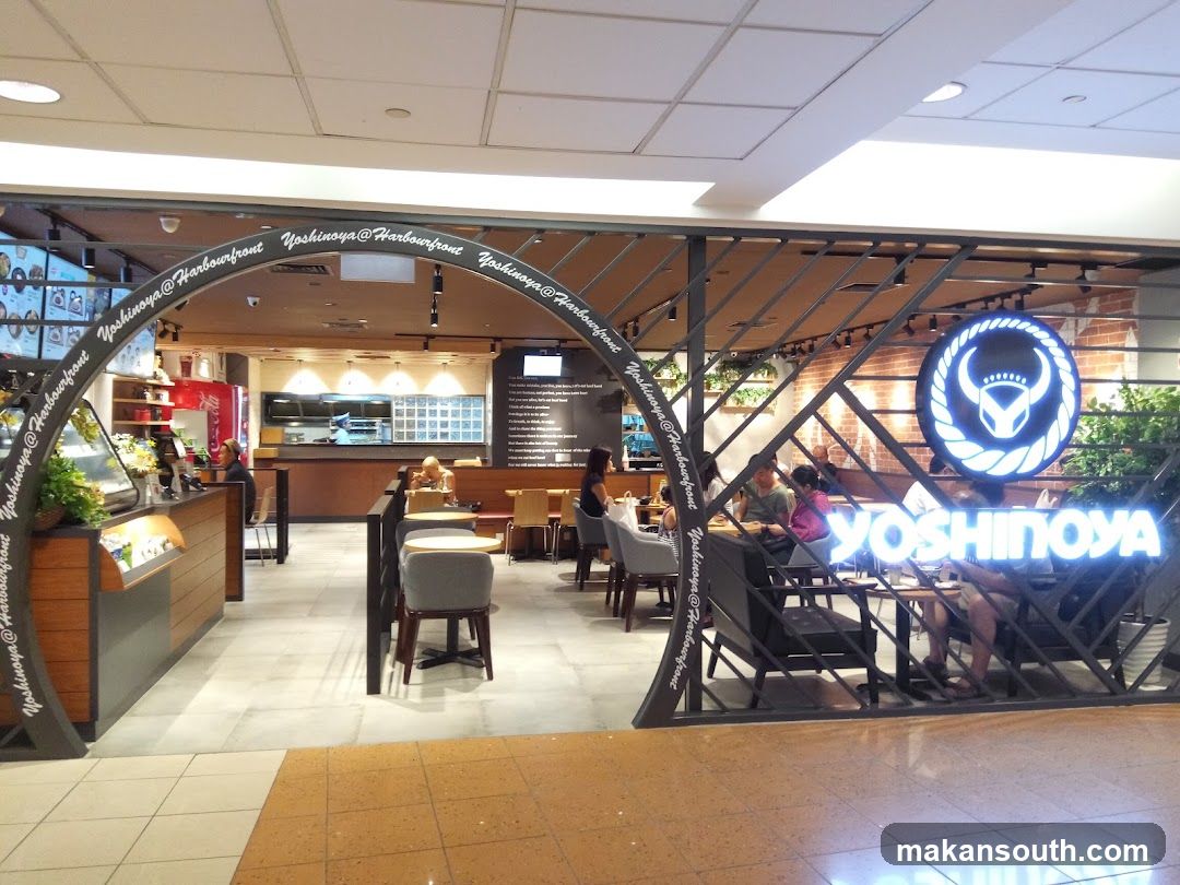 The exterior of a Yoshinoya restaurant with its prominent signage and logo. The restaurant features an arched entrance with the name "Yoshinoya@Harbourfront" repeated along the curve. Inside, customers are seated at tables, and the kitchen area is visible in the background.