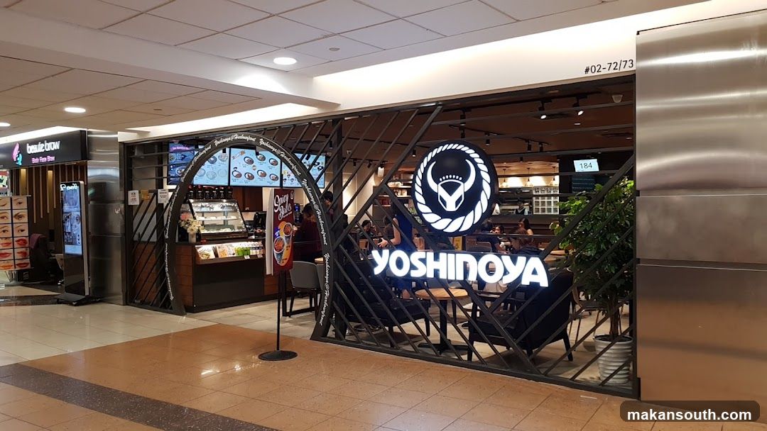 The exterior of a Yoshinoya restaurant with its prominent signage. Inside, customers are seated at tables. To the left, there is a display case with food items and a menu board above it.