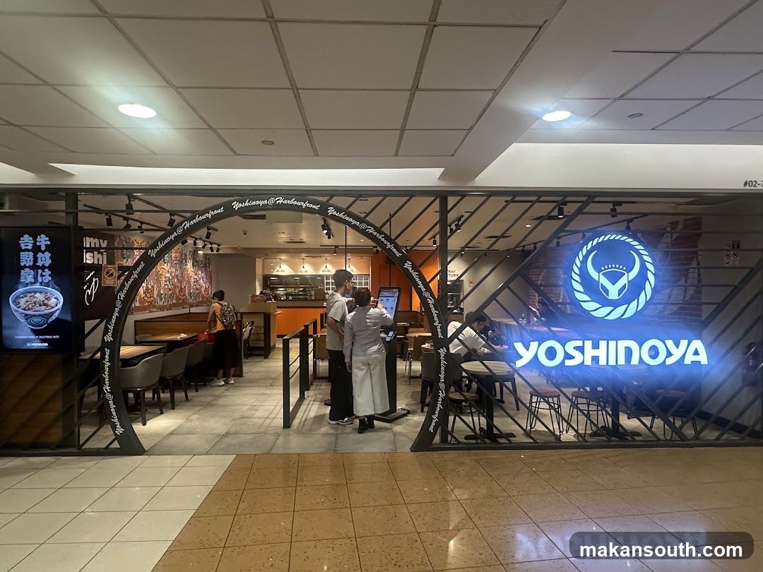 The exterior of a Yoshinoya restaurant. The entrance is framed by an archway with the restaurant's name repeated along its curve. A large, illuminated Yoshinoya logo and name are visible on the right side of the storefront. Inside, tables and chairs are set up, and staff members are visible behind the counter. A digital menu board is also present.