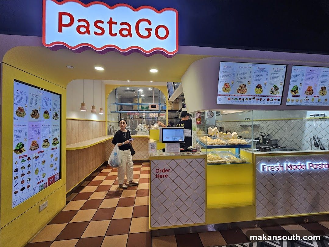 A brightly lit, modern pasta shop with a prominent "PastaGo" sign. The interior features yellow accents, checkered flooring, and digital menus displaying various pasta dishes. A woman stands near the counter, and staff are visible behind the service area. The phrase "Fresh Made Pasta" is illuminated on the counter.