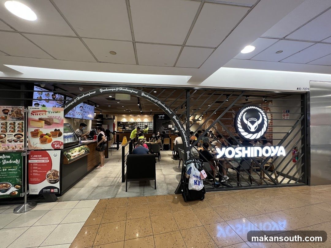 The exterior of a Yoshinoya restaurant with its prominent logo and name visible. The entrance is partially open, revealing the interior seating area and staff behind the counter. A menu board and promotional posters are displayed to the left of the entrance.