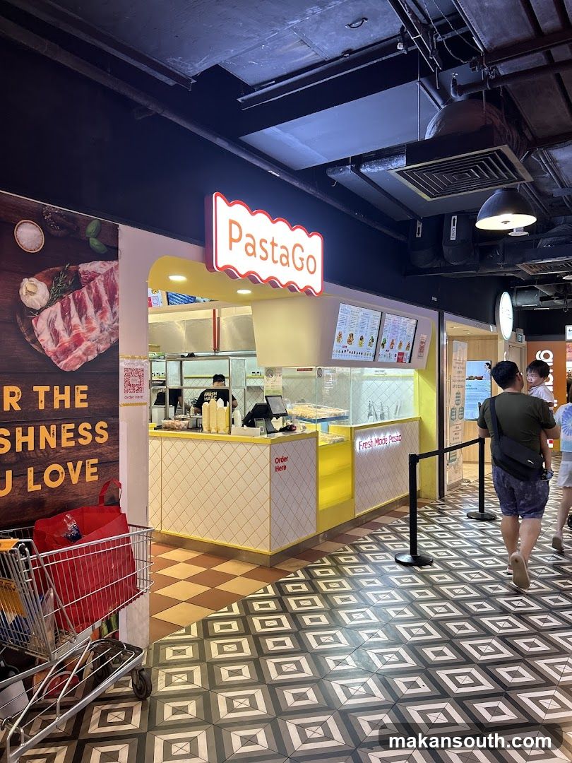 A brightly lit food stall with a prominent "PastaGo" sign. The stall features a yellow and white counter with a tiled facade. Behind the counter, staff members are visible preparing food. To the left of the stall, a large advertisement displays raw meat and the text "FOR THE FRESHNESS YOU LOVE". A shopping cart with a red bag is parked nearby. The floor has a distinctive geometric tile pattern.