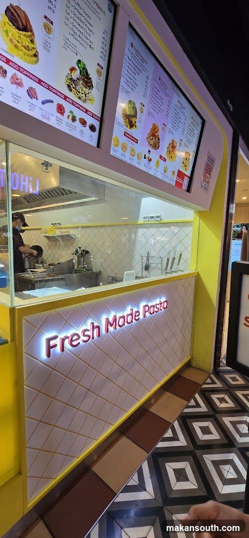 A brightly lit pasta shop with a yellow exterior and a tiled facade. The words "Fresh Made Pasta" are illuminated in pink lettering on the front. Behind a glass partition, a chef is visible preparing food in the kitchen. Digital menus displaying food items and prices are mounted above the counter.