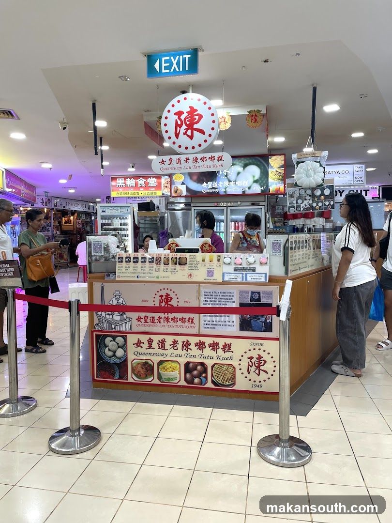 A food stall with a red velvet rope barrier in front. The stall has a sign with Chinese characters and English text that reads "Queensway Lau Tan Tutu Kueh". There are images of food items on the sign, including what appears to be tutu kueh, dumplings, and waffles. Several staff members are visible behind the counter, preparing food. Customers are waiting in line. An "EXIT" sign is visible overhead.