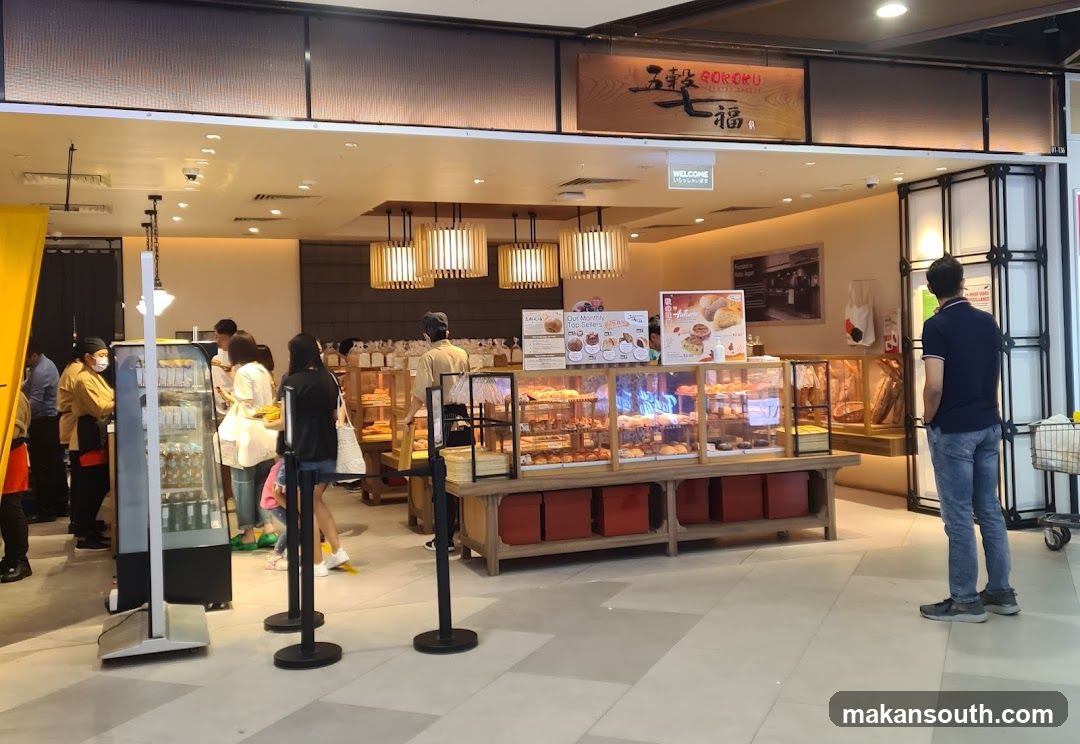 A Goroku bakery storefront with a display of baked goods. Customers are seen browsing and purchasing items. The bakery has a warm and inviting ambiance with wooden accents and soft lighting.