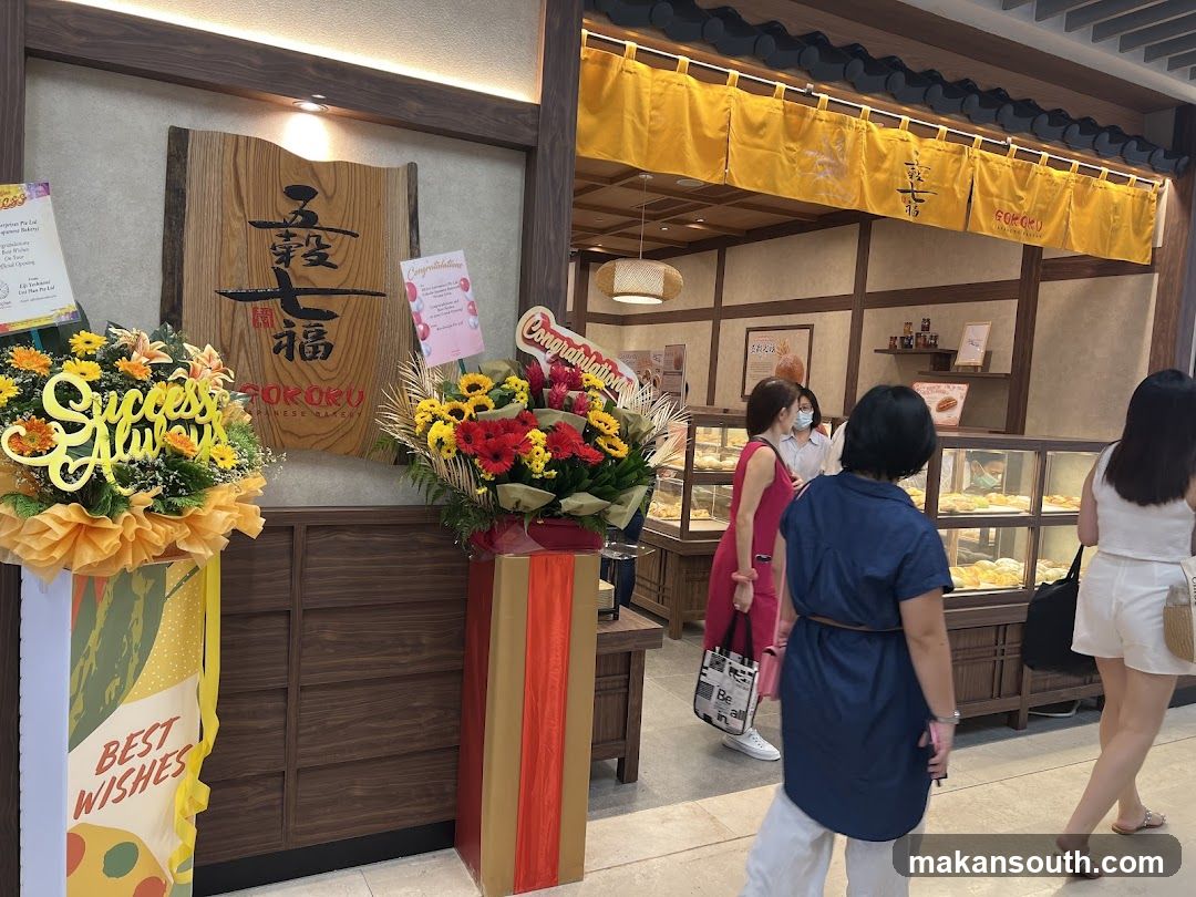 A wooden sign with Japanese characters and the name GOKOKU JAPANESE BAKERY is mounted on the wall. To the left, a floral arrangement with the words "Success Always" is displayed. To the right, another floral arrangement with a "Congratulations" banner is visible. The bakery interior features display cases filled with pastries and bread, with customers browsing. Yellow banners with Japanese text hang above the display area.