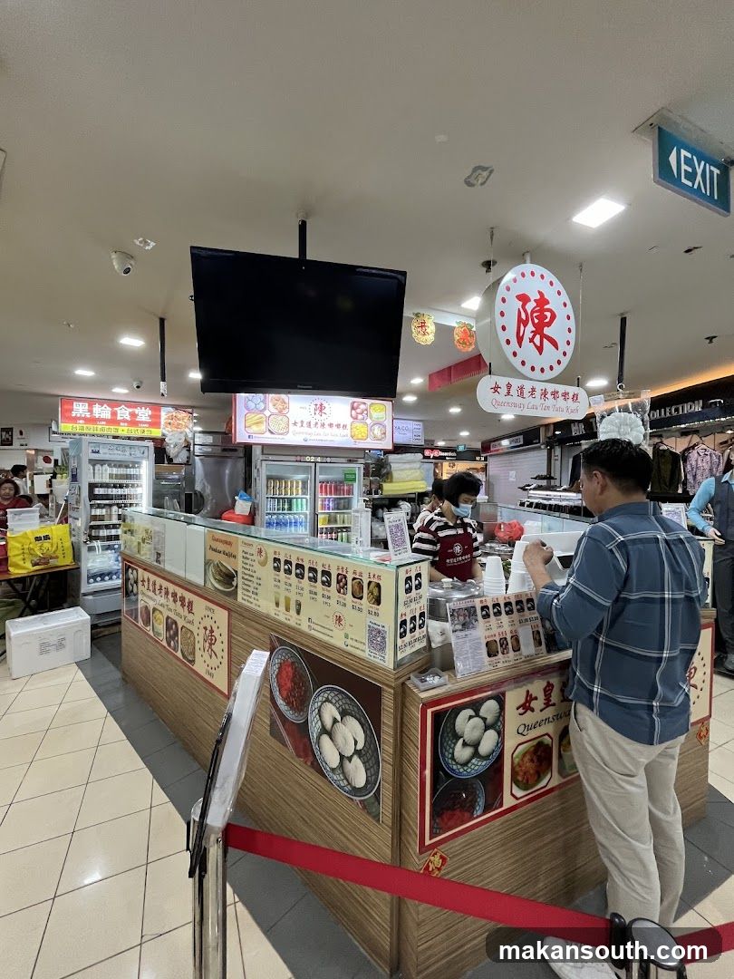 A food stall in a mall with a display of food items and a menu board. A customer is being served by a staff member.