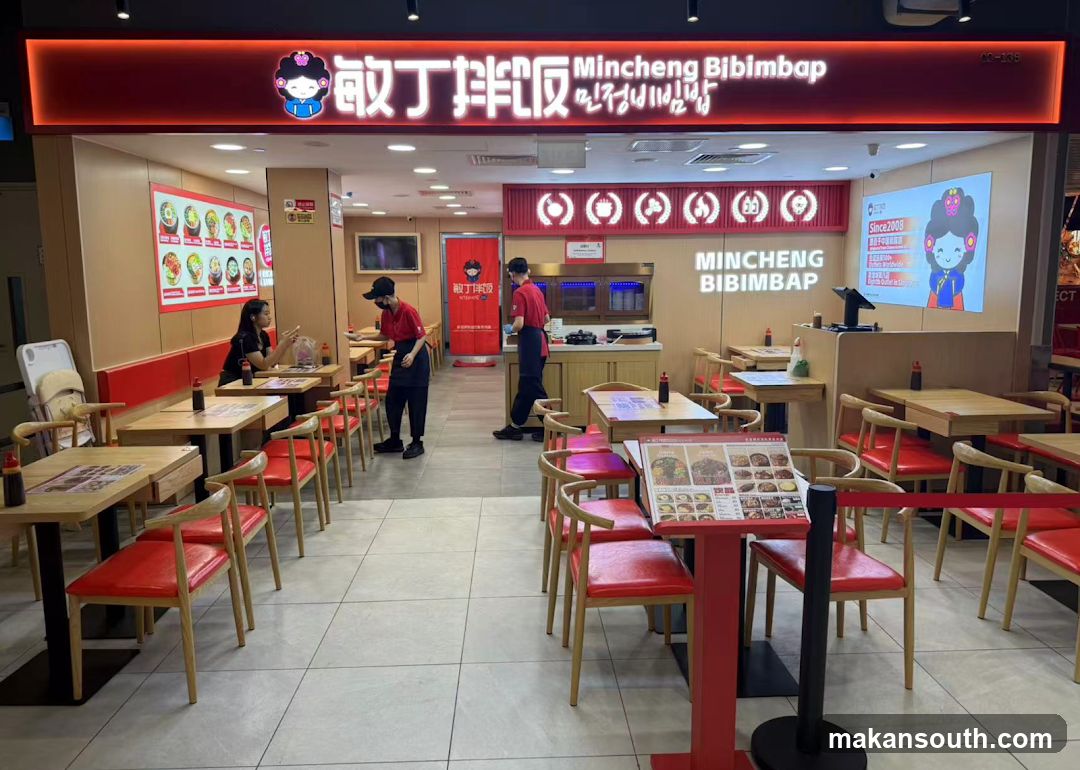 The exterior of a Mincheong Bibimbap restaurant. The storefront features a prominent red sign with the restaurant's name in English and Korean, along with a cartoon character. Inside, tables and chairs are visible, with some staff members attending to customers and preparing food. A menu board displaying various dishes is mounted on the wall.