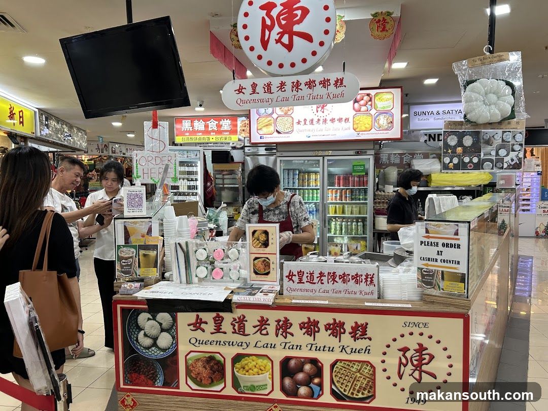 A food stall with a display of Tutu Kueh and other snacks. Customers are ordering at the counter.