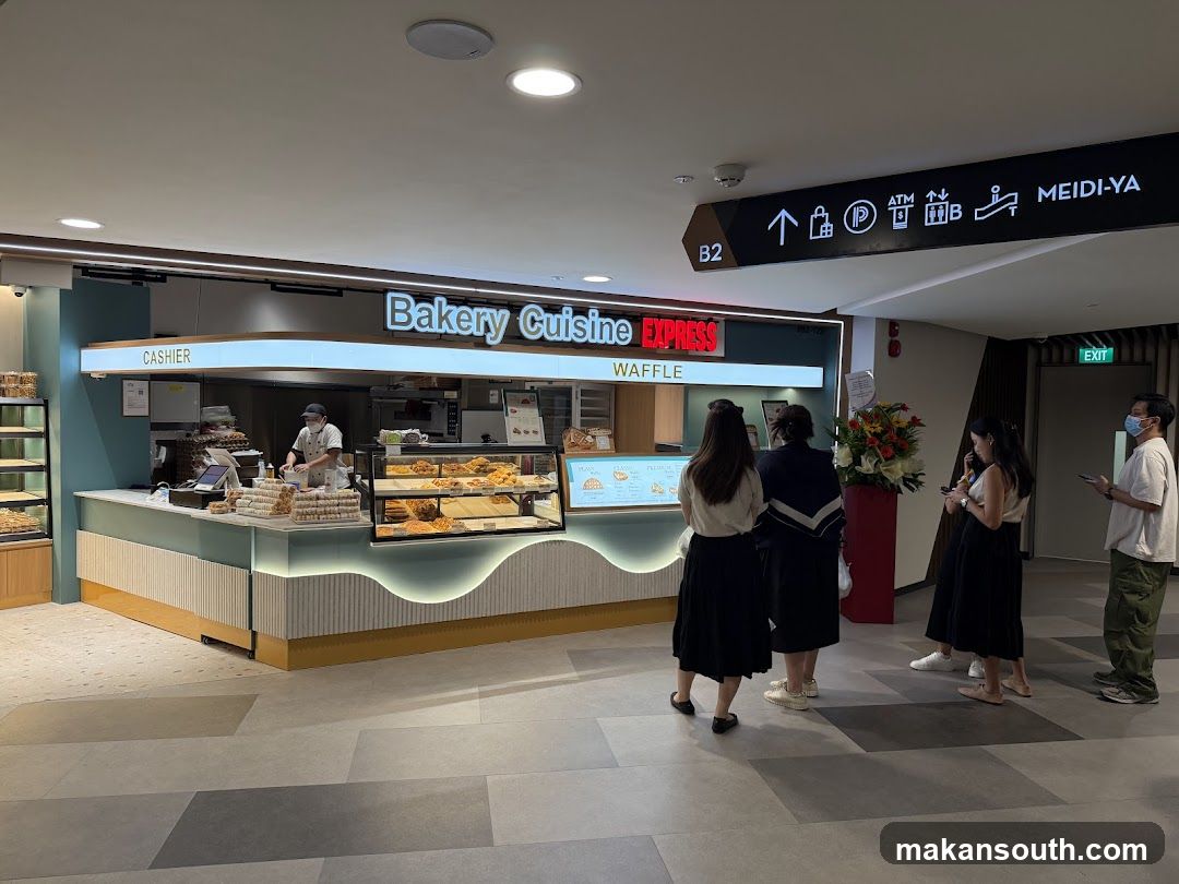 A bakery counter with a display of pastries and waffles. A cashier is visible behind the counter. Several customers are standing in line. Signage above the counter reads "Bakery Cuisine EXPRESS WAFFLE". A directional sign in the background indicates "MEIDI-YA" and amenities like ATM and restrooms.