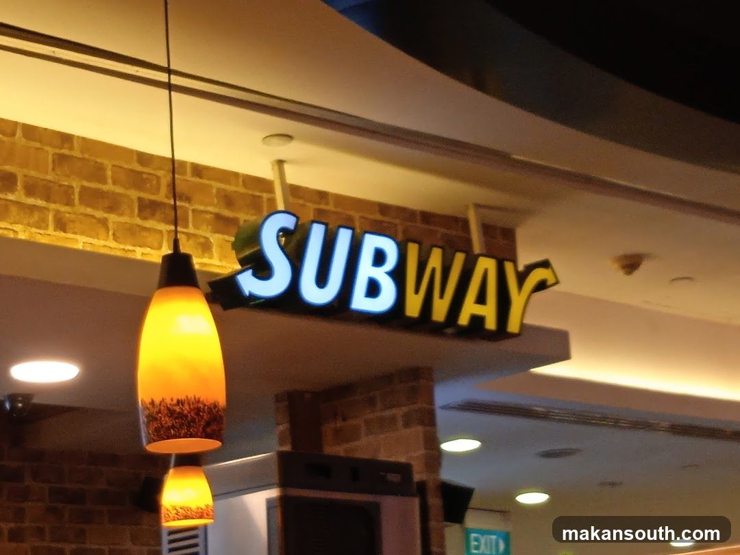 A Subway restaurant sign is visible, illuminated with white and yellow lettering. The sign is mounted above a brick-patterned wall. Two pendant lights with orange glass shades hang in the foreground.