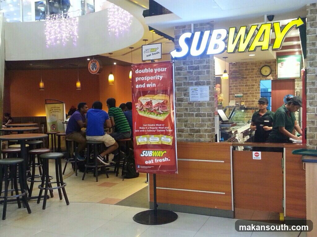 A Subway restaurant interior with customers and staff. The prominent Subway logo is visible above the counter. A promotional banner for a contest is displayed in front of the counter.